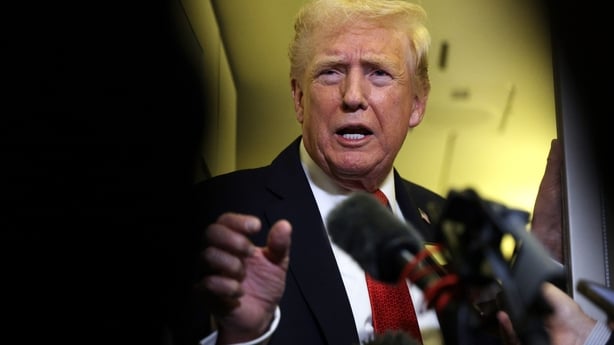 A head and shoulders image of US President Donald Trump wearing a white shirt, red tie and dark suit. He is looking ahead of the camera. There are two microphones in front of him. The background is white.