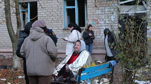 Several women wearing coats and hats are gathered outside a grey building that has been damaged. Its windows and door are broken.