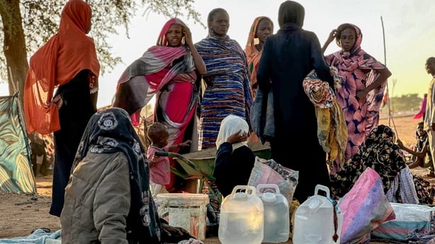 Several Sudanese women wearing brightly coloured clothing gather around white plastic containers of water while outdoors.