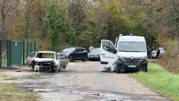 A burnt out car is seen on a road with other vehicles around it