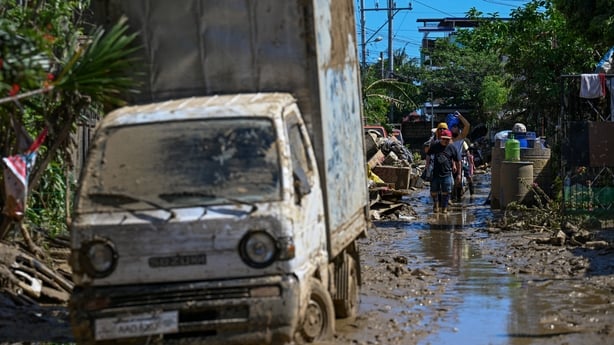 A white van covered in mud sits on a flooded street surrounded by rubble and debris.