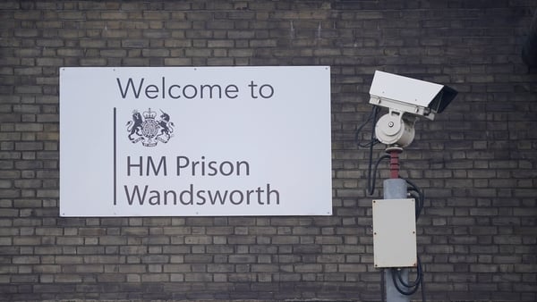 A white sign that reads 'Welcome to HM Prison Wandsworth' sits on a beige brick wall. A surveillance camera is beside it.