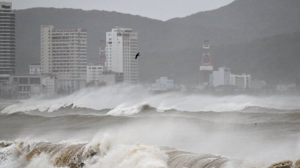 Strong waves crash ashore. White and grey buildings can be seen in the distance.