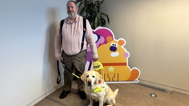 A guide dog and owner pose in an office in front of a cut-out and a small tree.