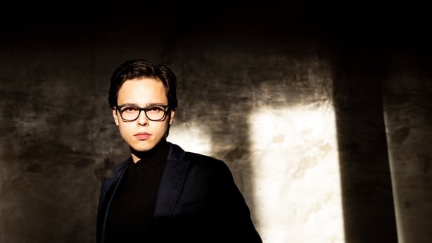 A photo of conductor Tarmo Peltokoski. He is standing looking at the camera, wearing all black, black glasses and has black hair. He has a serious expression. 