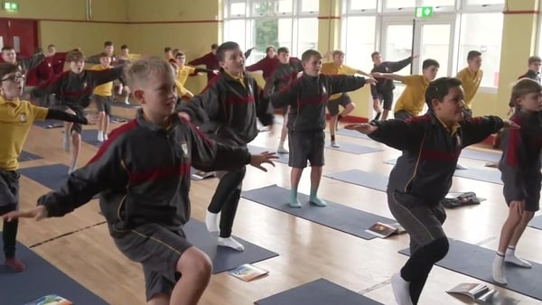 Young people in a school hall balance on one leg during a yoga lesson.