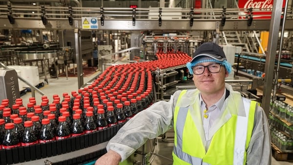 Man in a high-vis jacket and hair net pictured beside a Coca-Cola bottling conveyor belt
