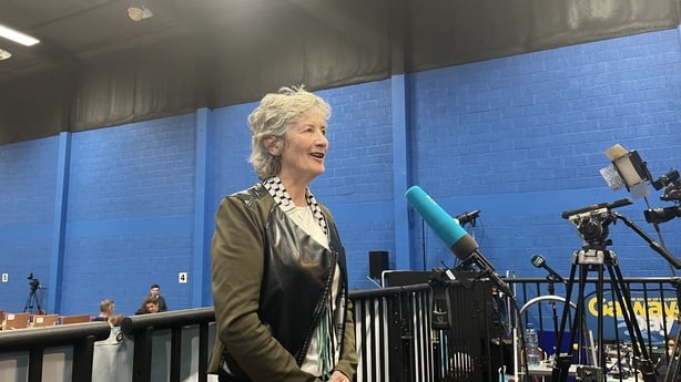 A woman with shoulder length grey hair wears a green and check jacket. She is facing a blue microphone. A blue wall is in the background