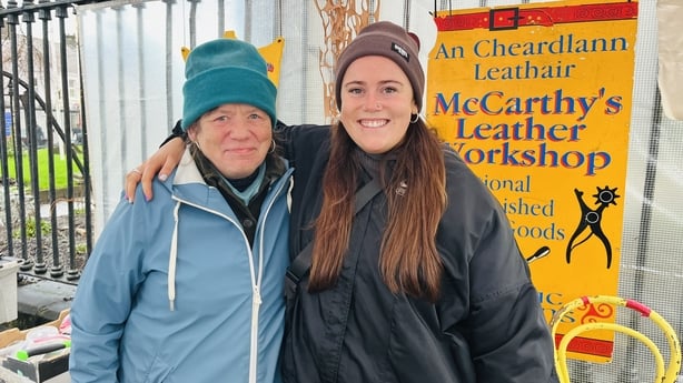 A young woman with long brown hair wearing a dark jacket and dark hat has her arm around an older woman wearing a blue jacket and blue hat.