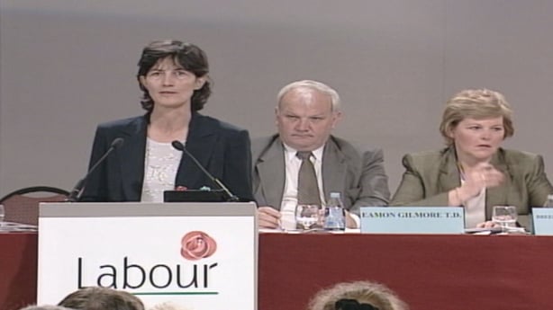 A woman with shoulder length dark hair wearing a white blouse and dark jacket stands in front of a table with a red table cloth. A sign reads 'Labour'.