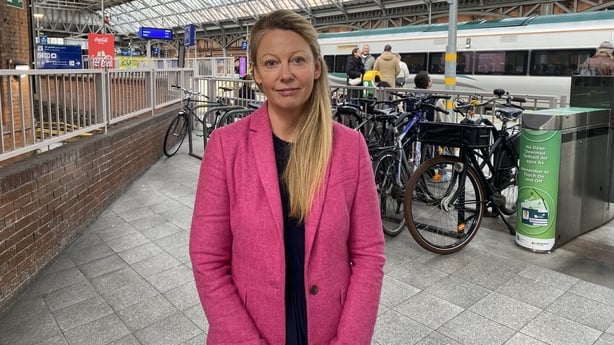 A woman with long blonde hair wears a black top and bright pink coat. She is inside a train station. A train and bicycles can be seen in the background.