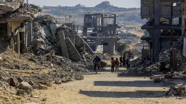Several people can be seen in the distance as they walk down a dirt track road surrounded by rubble and damaged buildings.