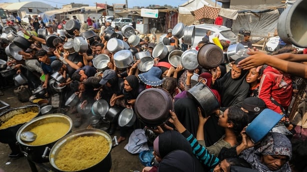 Dozens of people holding pots and pans gather closely together to try and get food from three large pots.