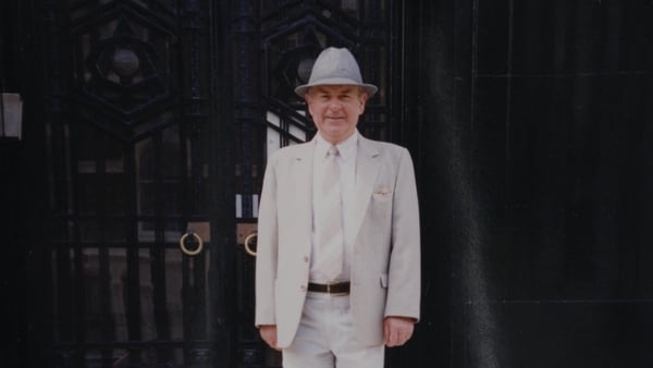 A man dressed in a grey suit and hat standing posing for a photo in front of a door