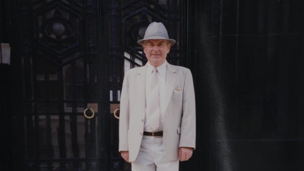 A man dressed in a grey suit and hat standing posing for a photo in front of a door