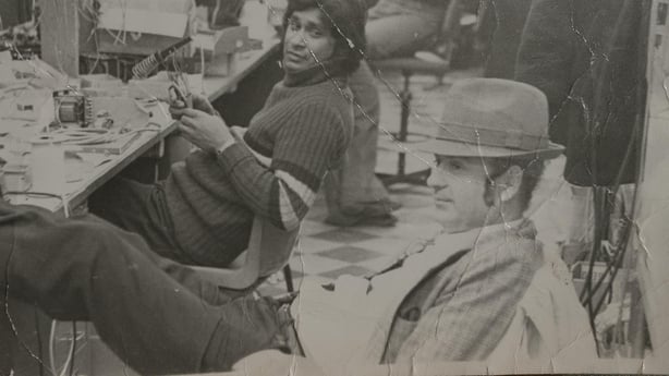 A black and white photo with two people - one wearing a distinctive hat with his legs up on a table
