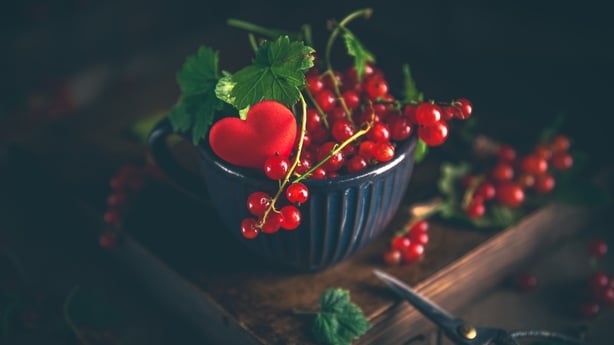 Freshly picked red currant berries and leaves are overflowing a green cup decorated with a red heart, sitting on a wooden box with gardening shears nearby