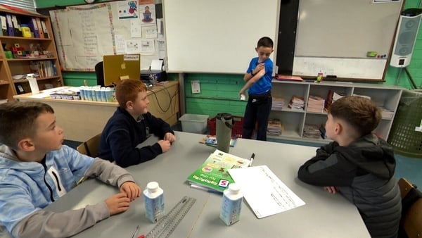 A boy stands up to do show and tell in a classroom in front of a table with three boys.