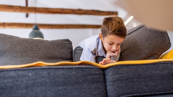 A young boy leans over a gray sofa, chin on hand, lost in thought in a cozy living room. Soft lighting and warm tones create a calm, reflective family moment.