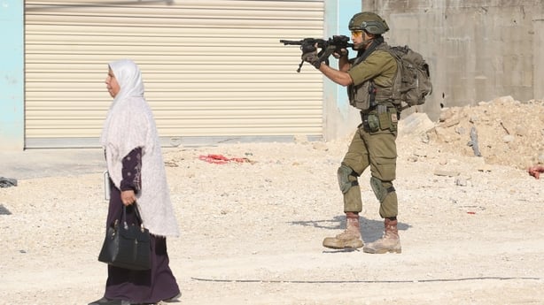 A woman wearing a white head cover and a black dress walks on a dirt path past a solider wearing green army clothing and holding a gun.