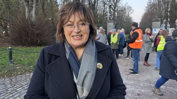 A head and shoulders image of a woman with shoulder length brown hair and glasses. She is wearing a dark coloured coat with a navy and light blue scarf. She is looking at the camera. She is standing outdoors with several people walking in the background.