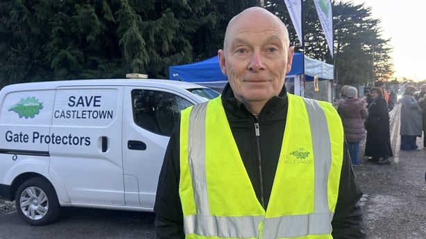 A head and shoulders image of a bald man wearing a black jumper and a high vis jacket. He is looking at the camera. A white van with the words 'Save Castletown' can be seen in the background, along with some trees and people.