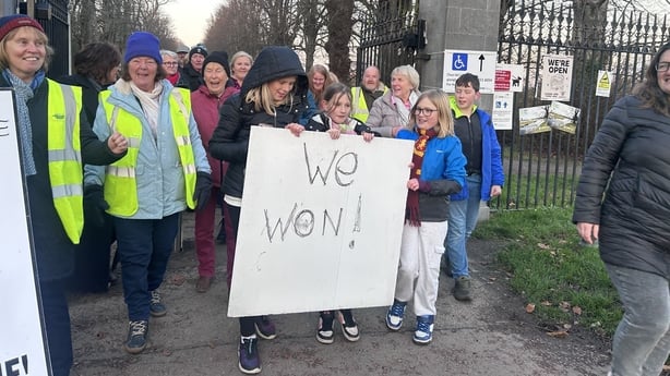 A group of people, some of whom are wearing high vis jackets, carry a large white poster that says 'We Won'. They are standing outside. There is a tall gate and some trees in the background.
