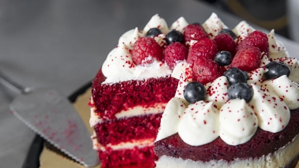 Red velvet cake decorated with raspberries, blueberries, and whipped cream is being sliced with a cake server