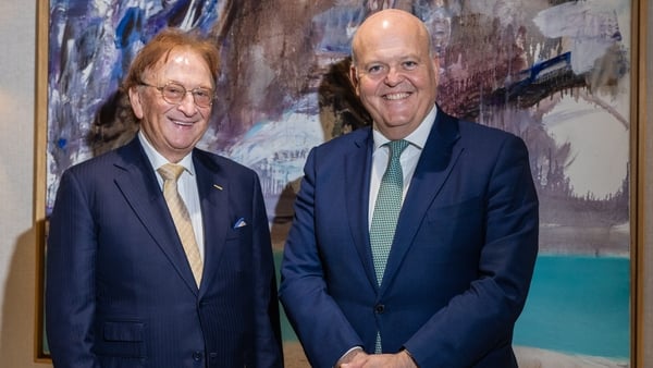 Two business men in suits pose for a photo with a painting in the background