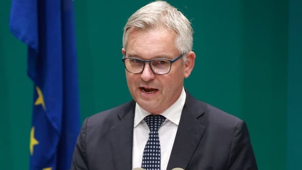 A head and shoulders image of a man with light grey hair. He is wearing a dark suit, dark tie and a white shirt. The background is green.