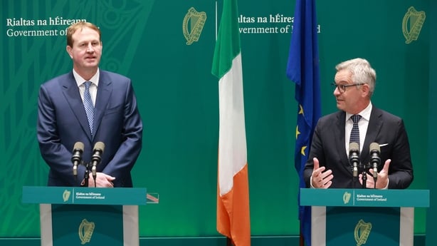 A tall man with light brown hair stands at a podium with two microphones. He is wearing a navy suit and tie and a white shirt. A shorter man with light grey hair stands beside him at a similar podium. He is wearing a dark suit and tie and a white shirt. The background is green.