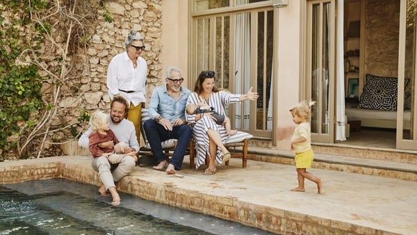 Wide shot of smiling grandparents relaxing with children and grandchildren beside pool at tropical villa during multigenerational family vacation