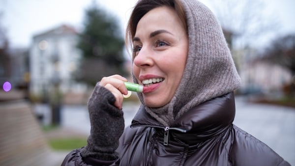 A woman applies lip balm while outdoors on a chilly day. She's wearing a dark brown hooded jacket and gray gloves.
