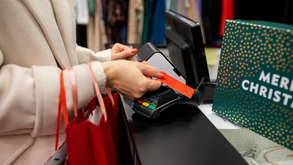 A shopper holds a red gift tag near a card payment terminal at a festive checkout, with a bright Merry Christmas sign and red gift bags, capturing holiday shopping and celebration.