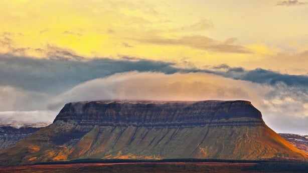 The iconic flat-topped Benbulben mountain in County Sligo rises dramatically from the Irish landscape, crowned by smooth lenticular clouds that form a natural cap above the limestone plateau during golden hour. This legendary Irish landmark represents both the country's unique geological heritage an