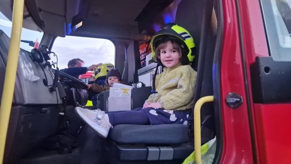 A small girl wearing a fireman's helmet sits in the cab of a fire truck with another child looking on