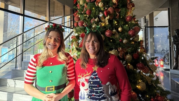 Two women pose in front of a Christmas tree wearing festive jumpers.
