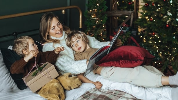 Happy mother with two sons siblings opening Christmas gifts at Christmas morning. Beautiful mother in white wool sweater and children reading book at christmas on the bed.