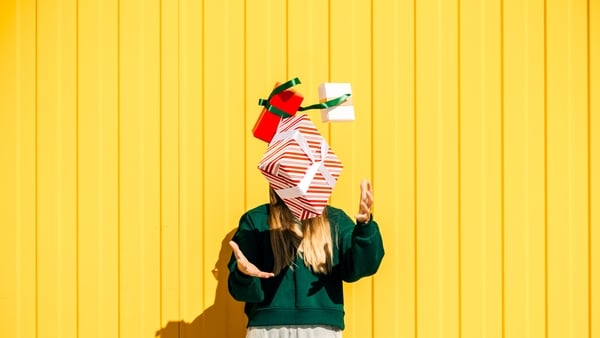 Woman in Christmas hat and green sweater catching Christmas gift boxes against the yellow wall.