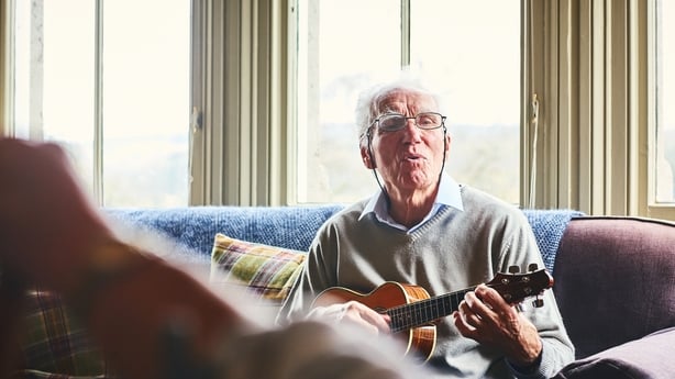 Happy senior man singing song and playing guitar for a woman sitting in front at home