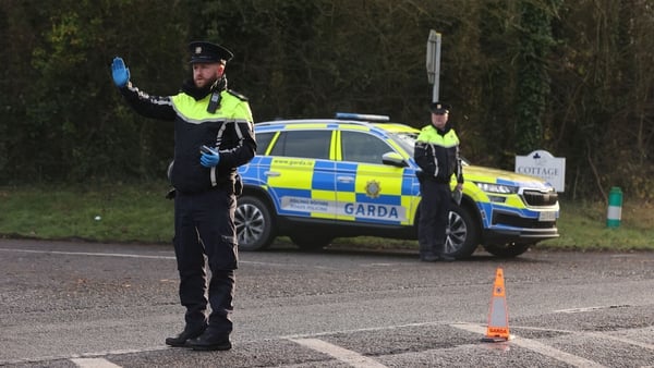 A garda raises his hand at a checkpoint with a garda car and a second garda in the background