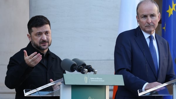 A man with short dark hair stands at a podium with two microphones. He is wearing dark clothes. A taller balding man stands at a podium beside him. He is wearing a navy suit and tie with a white shirt. The background is white.