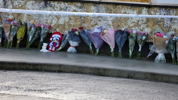 Dozens of bouquets of flowers stand in a row against a fire damaged wall. A garda no entry tape hangs above them.
