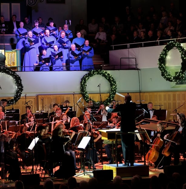the Palestrina Choir in blue robes, in balcony with RTÉ Concert Orchestra on stage at Sunday Miscellany Christmas concert, National Concert Hall, Dec 2025