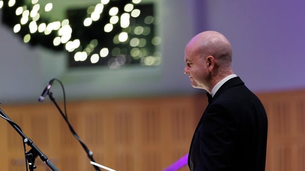 conductor Gavin Maloney standing at podium before the Orchestra, in dark stage light, for Sunday Miscellany Christmas concert, National Concert Hall, 2025 