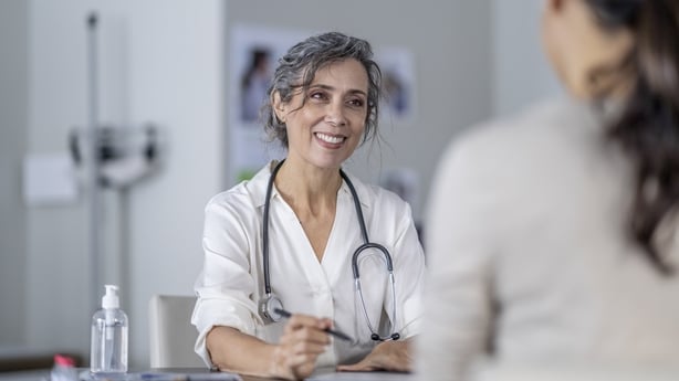 A mature female doctor points to the screen of her tablet laying out on the table in front of her as she shares her patients test results with her. She is wearing a white lab coat and has a stethoscope around her neck as she talks with the female patient of Asian decent.