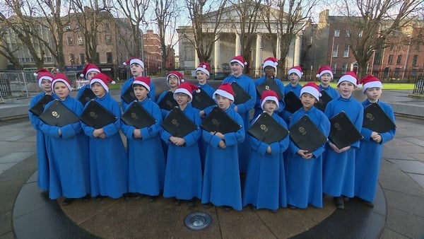 A schoolboy choir in blue robes wearing Santa Hats in a public park