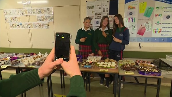 Three girls pose at a bake sale in their school as a person out of shot takes a pic on a phone.