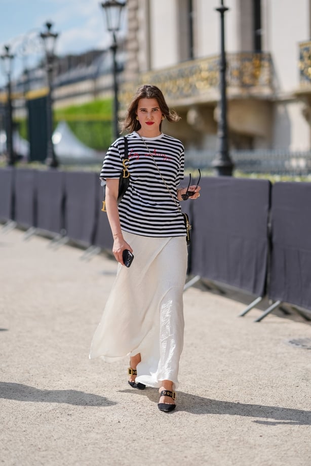 A guest wears gold earrings, black and white striped oversized short sleeve shirt with red print, black sunglasses in hand, a gold ring, shiny black Dior Saddle bag, shiny black leather bag with gold chain, gold bracelet, white flowy pattern maxi skirt, black with gold and black leather belt pointed