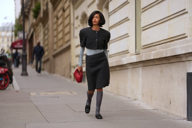 A guest wears a red quilted leather clutch bag from Miu Miu, a charcoal grey cropped wool jacket with short structured sleeves and two large front buttons layered, a light grey ribbed long-sleeve knit top, a dark charcoal grey wool knee-length skirt, knee-high grey ribbed socks scrunched at the cal